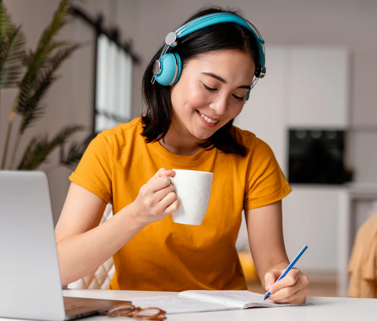 A learner in headphones taking notes beside a laptop