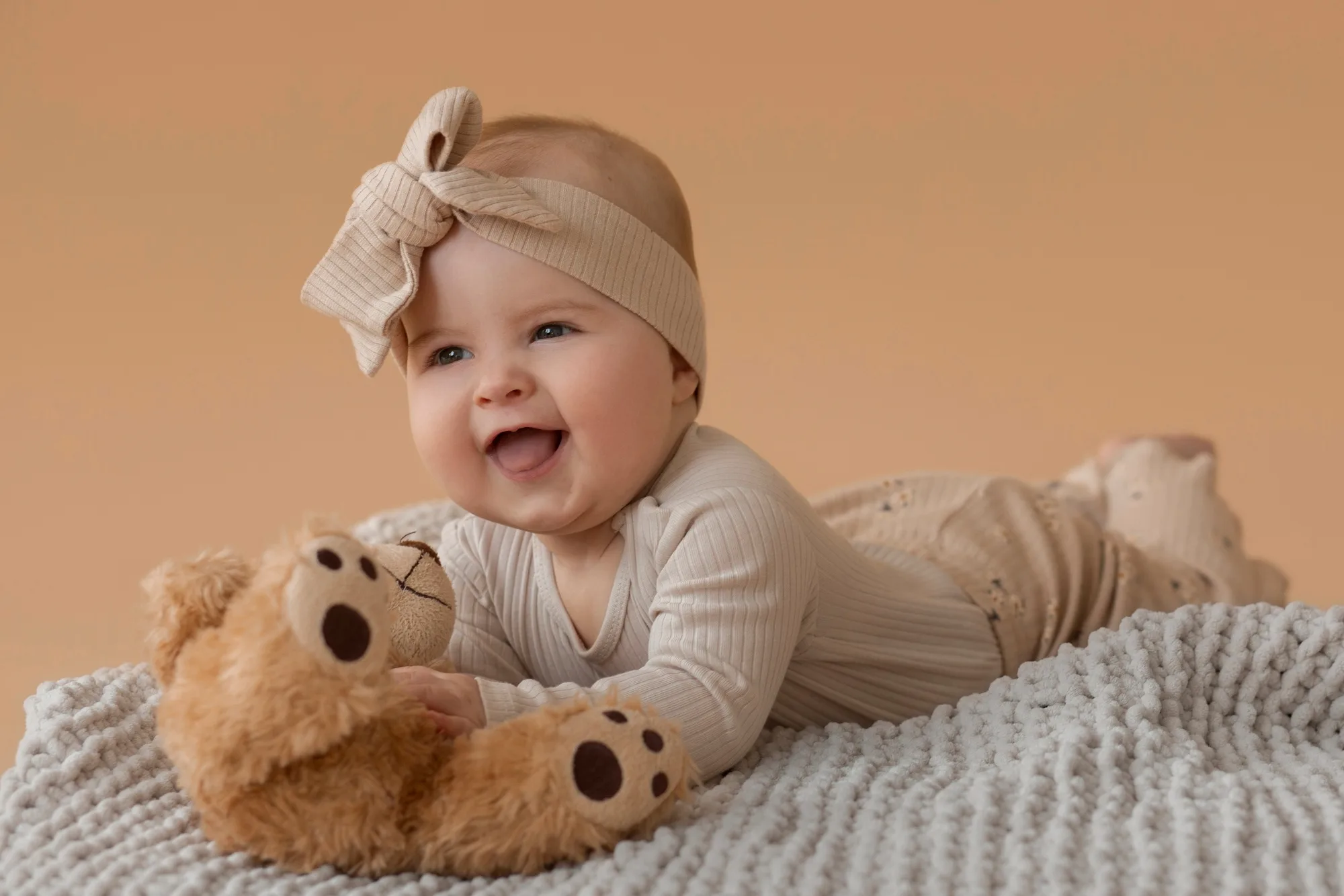 Smiling baby in a cream bow headband lying on a knitted blanket beside a teddy bear