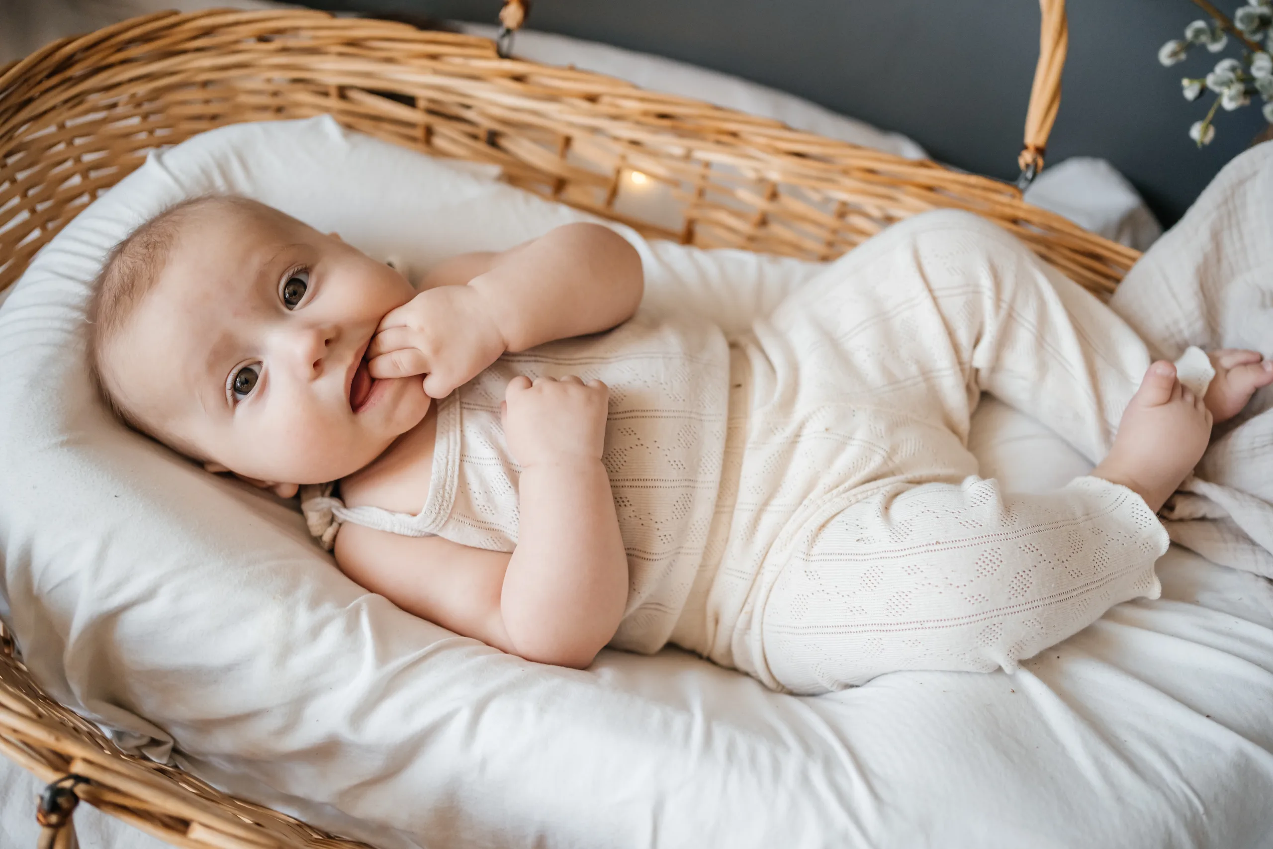 Baby in a cream romper lying in a woven bassinet with a hand at their mouth
