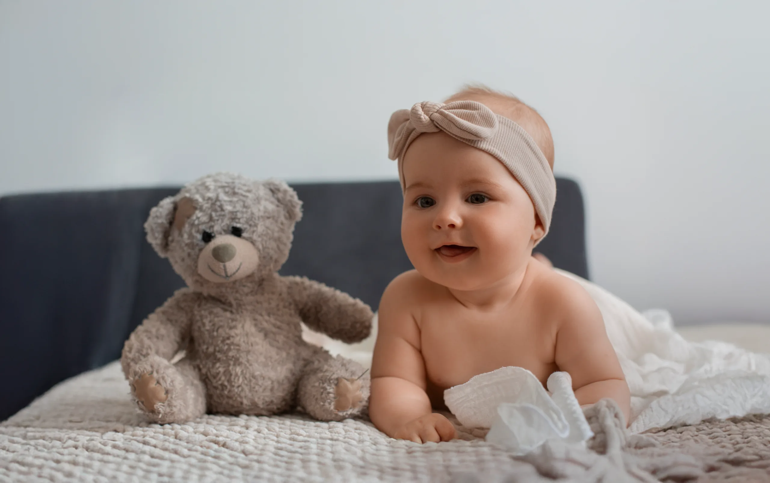 Baby in a bow headband sitting on a bed next to a grey teddy bear