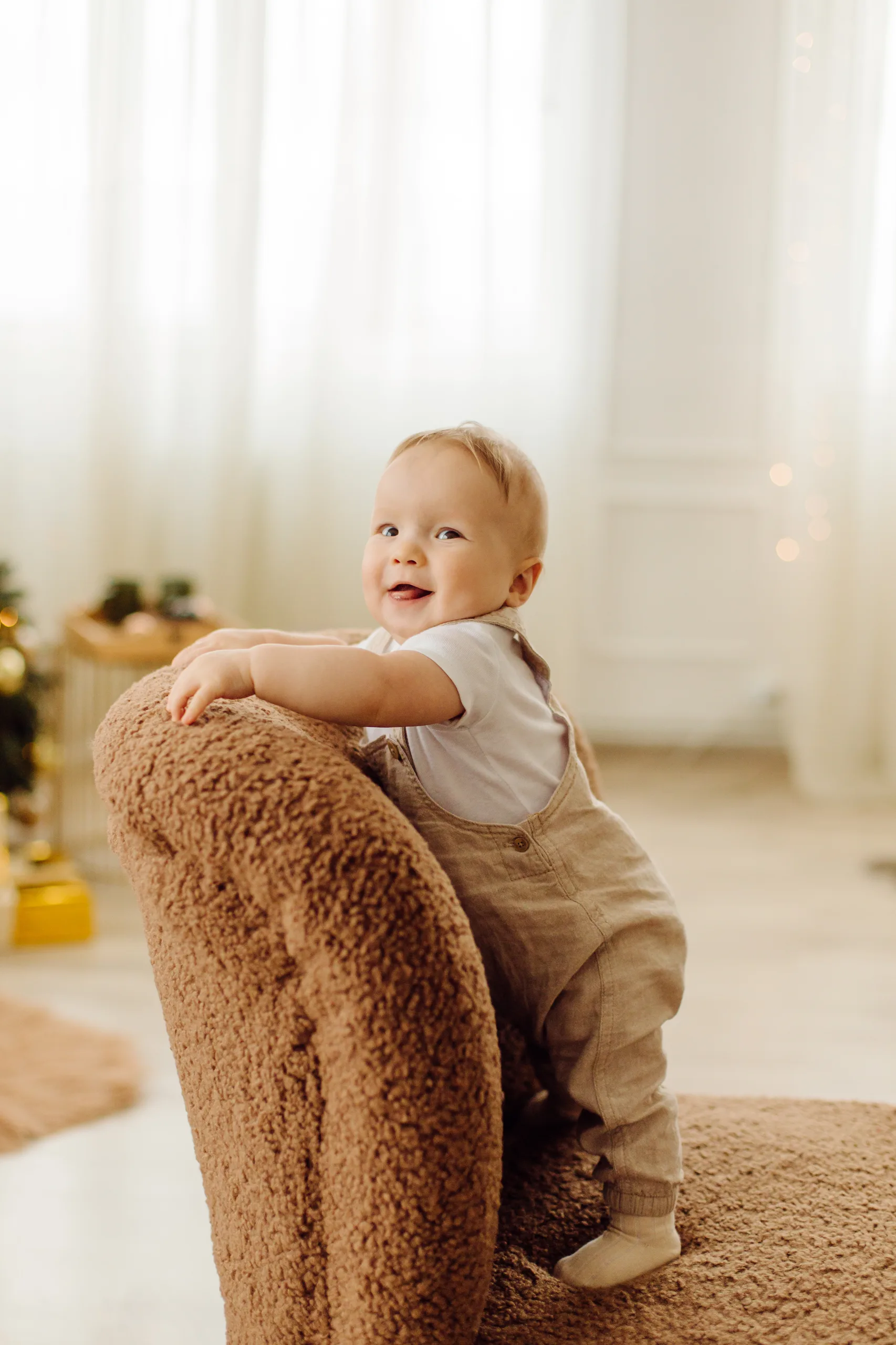 Baby in a linen dungaree set leaning on a plush chair