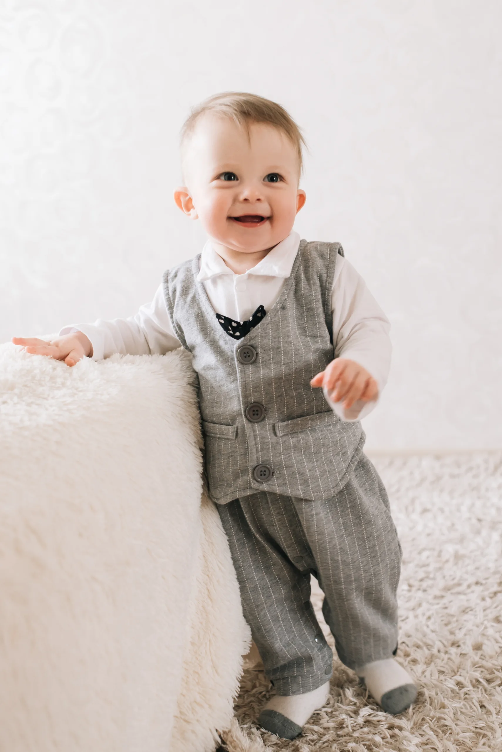 Baby in a grey waistcoat and shorts standing by a sofa