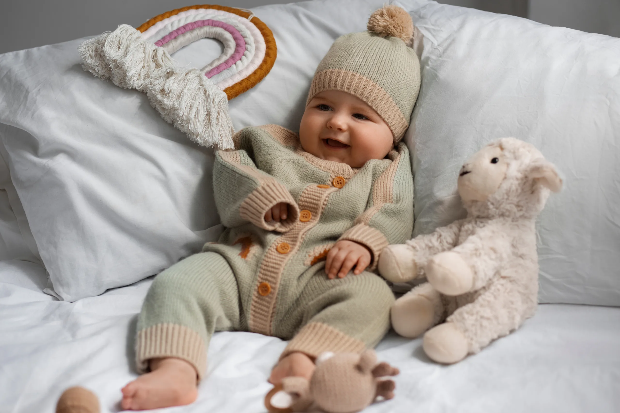 Smiling baby in a knit outfit and bobble hat lying on a bed beside a plush lamb and a macrame rainbow