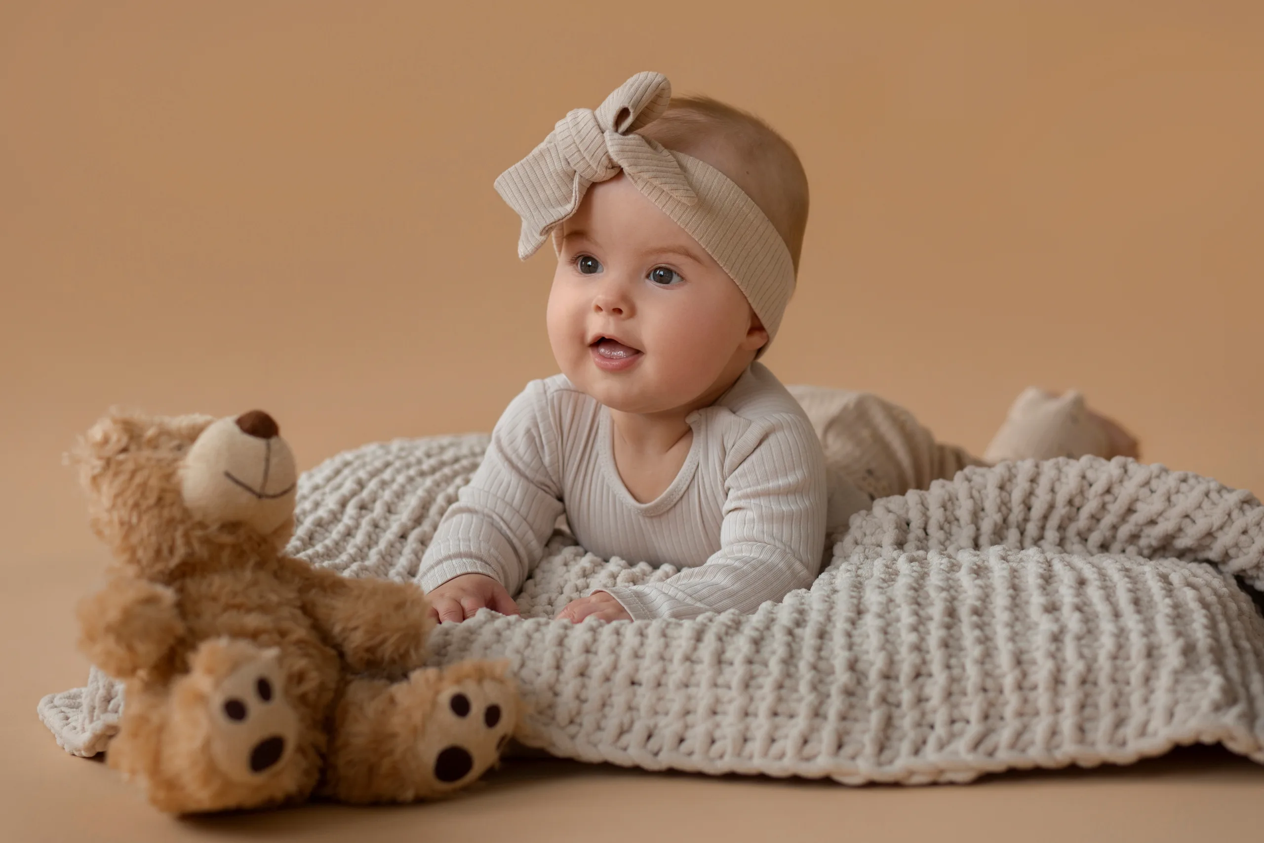 Baby in a cream ribbed outfit and matching bow headband lying on a chunky knit blanket beside a teddy bear