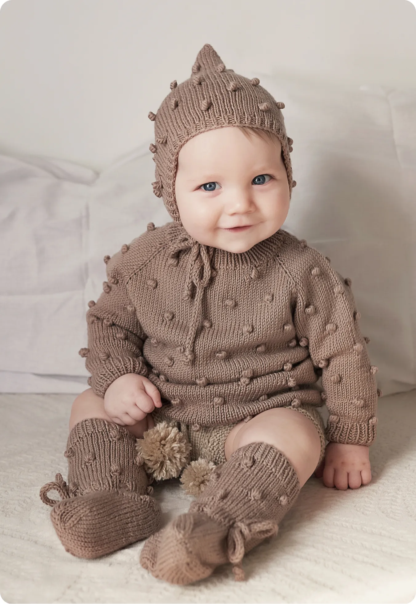 Smiling toddler in a knitted bobble bonnet and romper sitting on a cream bed