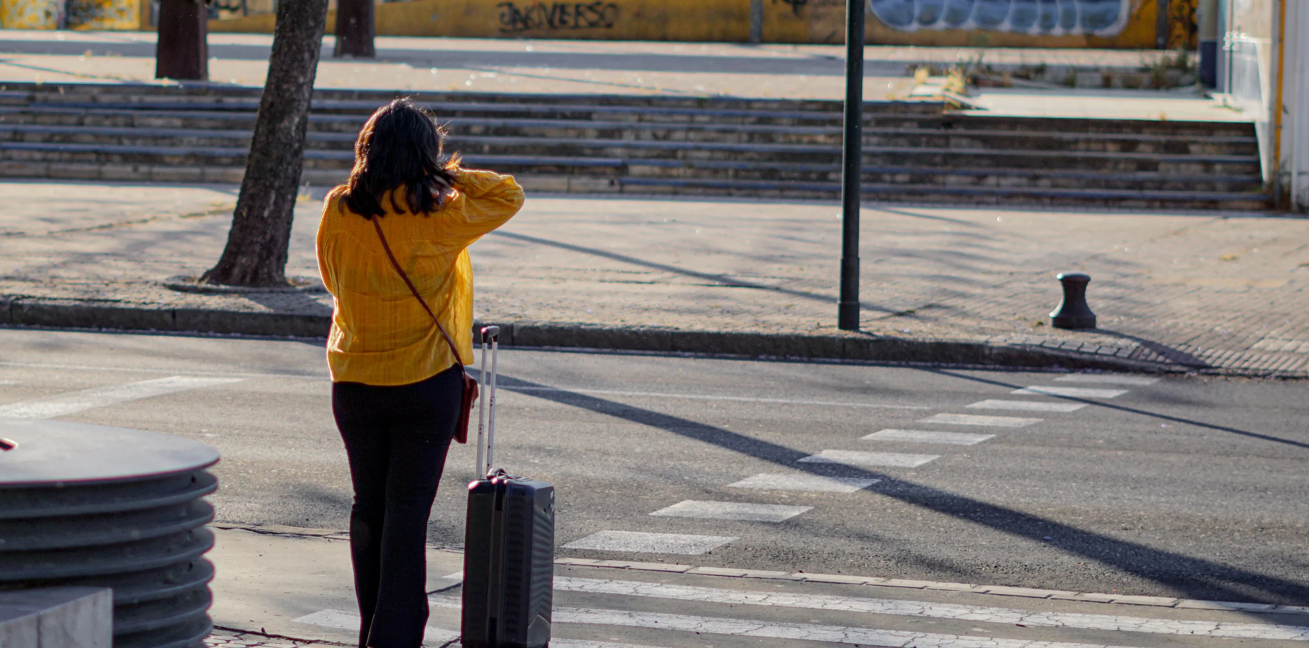 Traveller in a yellow jacket waiting at a city crossing with a wheeled suitcase
