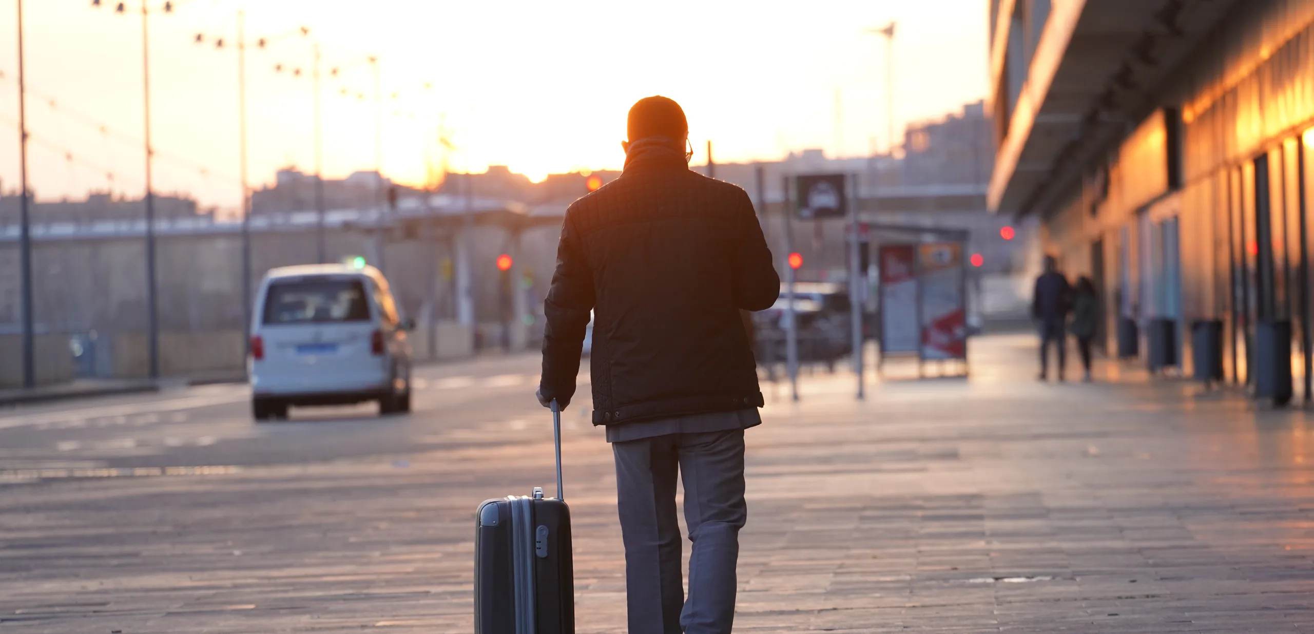 A traveller wheeling a suitcase across a station forecourt at sunrise