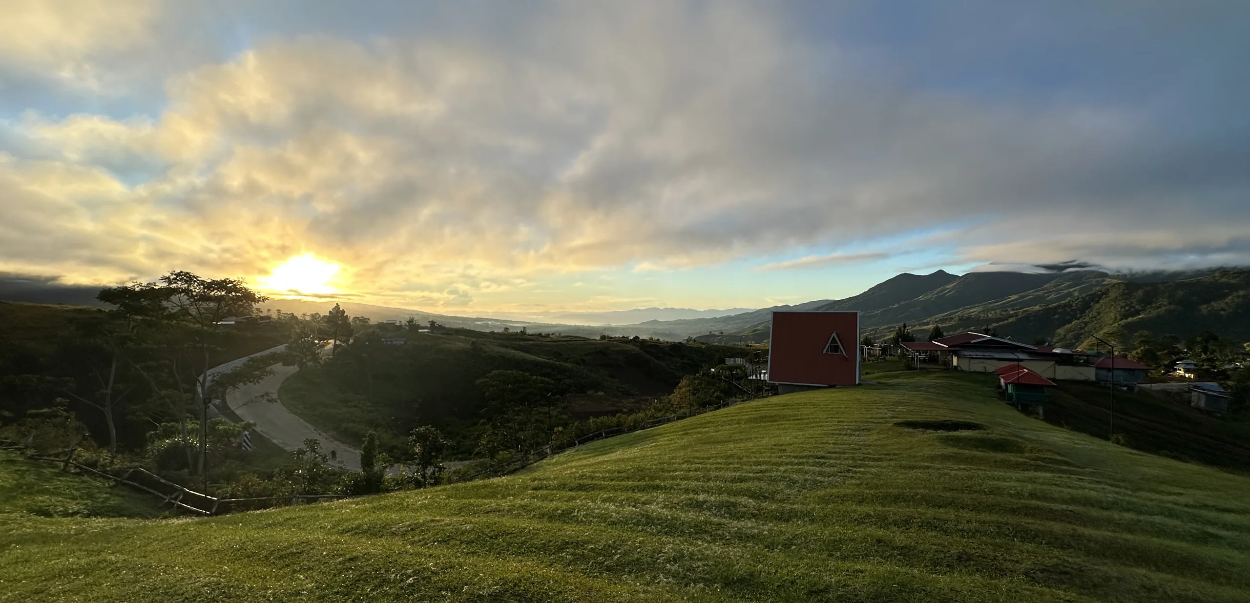 Sunrise over green hills with a red A-frame cabin and distant mountains